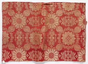  An intricately patterned textile or tapestry displaying a symmetrical floral design in shades of crimson red, pink, and beige with large central flowers interconnected by smaller blossoms and foliage details.