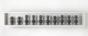  A collection of cylindrical chess-like pieces displayed in a long, shallow, light-grey display case, arranged in a straight line and varying in design, all rendered in a monochromatic color scheme.