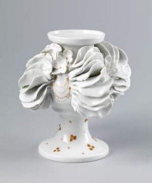  An ornate white porcelain vase or candle holder with a detailed ring of sculpted flowers resembling roses at its center, a smooth cylindrical neck and base, accented with small flecks of gold.