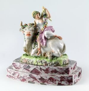  A porcelain figurine depicting a young woman with a green floral wreath in her hair, wearing a pink top and a white dress with blue and gold detailing, gently touching the head of a bull with a garland of flowers around its neck. The bull is mottled white and light brown, with a small white goat climbing on it. The figures stand on a textured green base simulating grass with small flowers, all atop a marbled pink, white, and gray base.