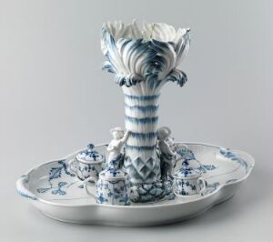  A blue and white ceramic centerpiece with Delftware-style designs, featuring a vase with a ruffled, flower-like top centered on a decorative plate adorned with intricate pastoral scenes.