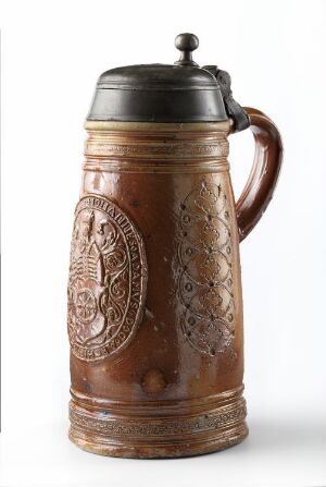  A traditional glazed ceramic German beer stein with detailed relief decorations and a pewter lid. The stein is predominantly earthy brown with lighter and darker accents, photographed against an off-white background. Artist name and title are unknown.