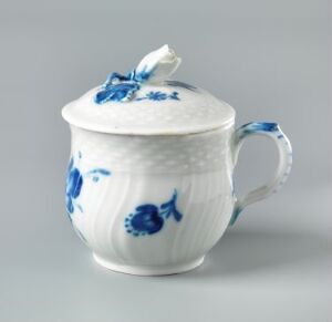  A white porcelain mustache cup with a ribbed texture and dark blue floral patterns, featuring a handle and a lid with a blue petal design on the top, designed to elegantly protect a mustache while drinking.