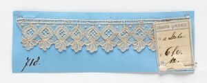  A piece of white lace trim with a diamond and floral pattern, displayed on a light blue paper background alongside a vintage paper tag with printed and handwritten text, suggesting a cataloging or sales sample.