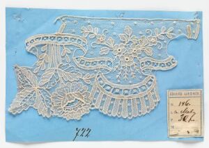  Intricate white lace with a floral pattern displayed on a pale blue background, with a handwritten label to the right, indicating catalog or reference information.