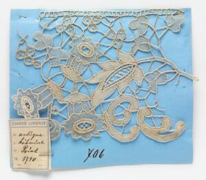  An intricate beige paper cutout with floral and foliate designs, displayed on a pale blue background, with a small handwritten label in the corner showing the number "906."