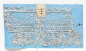  A piece of intricate light blue lace with a floral pattern and scalloped edge is laid out on a slightly lighter blue background, with a small handwritten tag affixed to it and a catalog number in the corner.