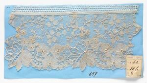  Piece of white or cream lace fabric with a floral pattern and gold threading details laid out on a powder blue background, featuring an ornamental border and an aged paper tag with handwriting in the lower right corner. The artist's name and title are unknown.