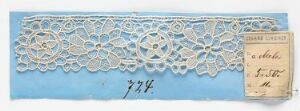  A strip of white lace with intricate patterns laid on a pastel blue background, accompanied by a vintage handwritten label and a bold number written beneath the lace. Artist name and title unknown.