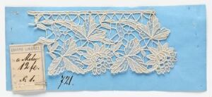  A delicate white paper lace artwork with a botanical pattern is displayed on a medium-blue textured paper background, accompanied by a small, aged paper tag with handwritten text at the bottom left corner.