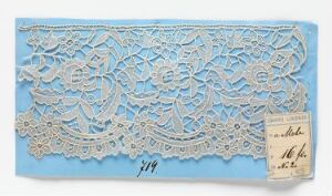  A piece of intricated white lace with floral patterns on a light blue paper background, accompanied by a handwritten tag on the right side. The artist's name and title are unknown.