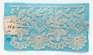  An ornate sky-blue textile with intricate white lacework displaying floral and geometric patterns, accompanied by a small, indistinct paper card on the left.