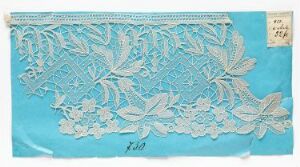  Light blue rectangular fabric with an intricate white lace floral pattern overlay, tapering upward from the bottom edge, with a small beige paper tag in the top-right corner. Artist name and title are unknown.