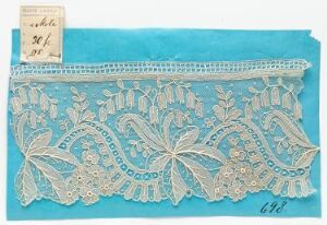  Antique floral-patterned lace with detailed eyelets and scalloped edges laid on a uniform light blue background, with a small, unlegible paper tag attached to the upper left corner and the number "646" handwritten below.