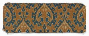  Elongated fragment of decorative patterned material with symmetrical fleur-de-lis or leaf-like motifs in mustard gold, deep teal blue, and slate blue on a textured golden background suggestive of aged parchment or woven fabric.