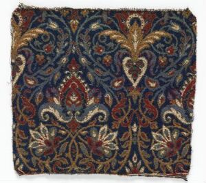  An ornate fabric fragment with a symmetrical pattern of paisley and floral motifs in rich colors of burgundy, gold, navy blue, forest green, and light blue, with frayed edges suggesting an antique or handcrafted textile.