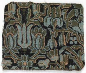  A piece of aged textile with a complex, symmetrical floral pattern in shades of teal, green, pale orange, cream, and brown, on a dark blue, almost black, background. The design is intricately woven, featuring large central flowers and surrounding foliate motifs with frayed edges suggestive of historical craftsmanship.