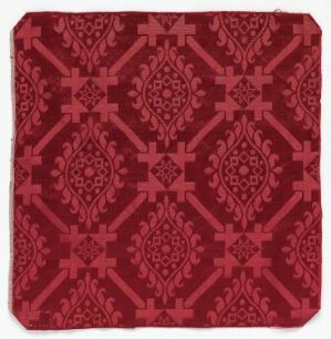  A square piece of fabric with an intricate, symmetrical pattern in various shades of red, featuring a combination of diamond shapes and curling, leaf-like designs, creating a repeating, ornate design that suggests historical use in home decor.