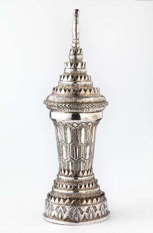  Ornate silver-colored decorative tower with intricate embossing featuring geometric and floral patterns, set against a plain, light background. The tower has a broad base, a detailed midsection, and a slender spire atop. The material reflects light, suggesting a metallic composition.