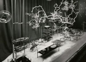  Black-and-white photo of an indoor exhibition featuring abstract suspended sculptures and organized display tables with various objects, against a backdrop of draped curtains, exemplifying modernist architectural photography.