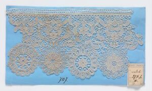  An intricately designed off-white lace with a detailed pattern of circular floral motifs and connecting meshwork is laying on a light pastel blue background, with a small, handwritten paper tag attached to its right side.