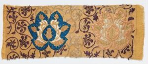  An antique textile piece with a faded beige background, featuring a central blue symmetrical emblem surrounded by brown and purple vine and floral motifs, showing signs of age with frayed edges.