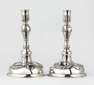  A pair of ornate silver candlesticks with embossed designs on their bases and gracefully curved stems, set against a light gray background.