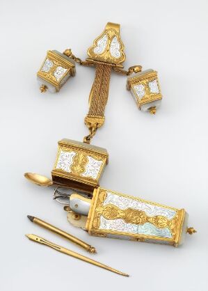  An ornate historical writing set with a gilded case featuring ivory inlay work, three smaller matching containers attached with a chain, and two writing instruments with golden handles displayed against a light-gray backdrop. Artistname and title: Unknown