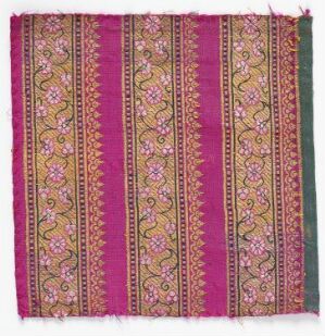  A square piece of richly designed textile with a dominant fuchsia pink background featuring intricate floral or paisley motifs in golden yellow, blue, white, and green. The fabric has purple and maroon borders with simple linked geometric patterns in yellow tones.