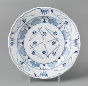  A round feldspar porcelain plate from the Blaaveis series by Gerhard Munthe, featuring a white background with hand-painted and stencil-painted underglaze decoration in various shades of blue. The center of the plate displays a cross-like pattern with floral motifs, surrounded by bands of stylized blue fish and wave patterns, all enclosed by a scalloped rim.