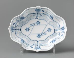  The image features a feldspar porcelain plate with hand-painted and stencil-painted blue floral underglaze decoration designed by Gerhard Munthe. The plate has a wavy edge and a central diamond motif surrounded by a chain of florals, showcasing a combination of traditional and modern porcelain decor on a white glossy surface.