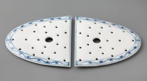  Two halves of a Svibelmønster feldspar porcelain platter with a white background, decorated with hand-painted underglaze blue leaf motifs and scattered black dots, each with a central hole, on a grey surface. Artist unknown.