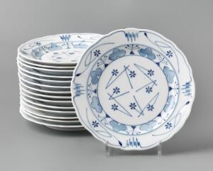  A set of "Blaaveis" feldspar porcelain plates, designed by Gerhard Munthe, featuring a central hand-painted blue and white snowflake-like motif, surrounded by smaller blue patterns and lined at the borders, displayed against a neutral gray background.