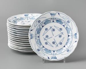  A stack of feldspar porcelain plates with intricate blue patterns on a white background, designed by Gerhard Munthe, titled "Blaaveis." One plate is displayed in the foreground, showcasing a starburst-like pattern in the center with a decorative border, suggesting a union of functionality and artistry in dining ware.