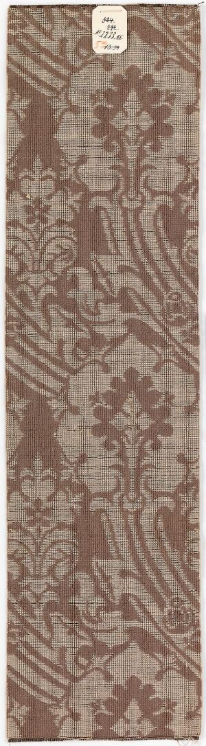  A narrow, vertical textile fragment with an elaborate pattern of stylized plant motifs in muted burgundy and dark brown, on a gradient beige background, suggesting an antique aesthetic.