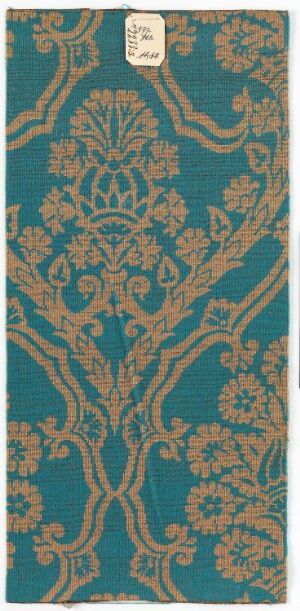  An ornate textile piece with a dark teal background, adorned with an intricate, symmetrical gold floral pattern. A white label with text is positioned near the top of the textile.