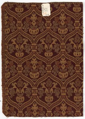  A segment of an ornate, dark and light brown patterned fabric or paper with a small white archival tag in the top left corner, featuring a detailed symmetrical design possibly used in historical bookbinding, wallpaper, or textile.