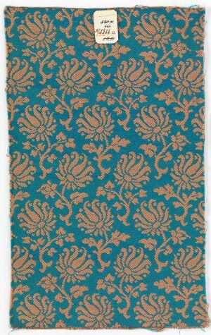  A detailed floral and plant motif pattern on a textile, with a turquoise background and ornate designs in shades of orange, gold, and beige. A small, rectangular label is affixed to the top of the fabric.