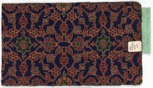  A detailed, ornate patterned fabric with a navy blue background featuring intricate floral and geometric designs in red, orange, green, and yellow, with a plain teal block on the left edge and a small white barcode label on the bottom right.