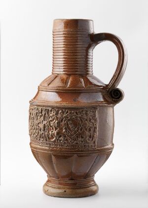  A ceramic pitcher with a warm brown hue and intricate etched patterns. The body is stout with vertical ribbing and a decorative band, while the flared lip and twisted handle add elegance to its historical design. The pitcher is set against a neutral light grey background.