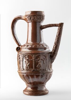  An ornate, glossy brown ceramic jug with a curved handle and detailed relief work depicting classical scenes, set against a solid, neutral background.