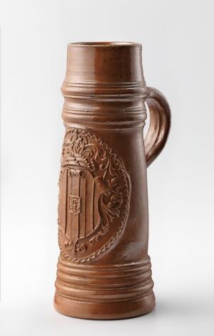  A glazed stoneware jug in warm brown, with relief decoration, made by Hubert Schiffer, illustrating a cylindrical form that tapers towards the top, a handle on the side, and an embossed oval medallion with a traditional scene.