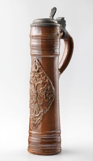  A traditional ceramic beer stein with a glazed brown surface and an ornate raised relief design on the side, featuring a metal lid with a thumb lever.