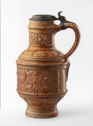  An ornate ceramic jug with a dark flip-top lid, a detailed bas-relief design featuring figures and foliage in shades of brown, and a sturdy handle, set against a plain white background.