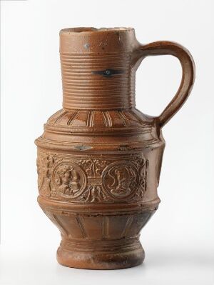 
 A traditional terracotta ceramic jug with a rounded body featuring ornate, embossed patterns, a ridged neck, and a single curved handle, set against a plain white background. Artist name and title are unknown.