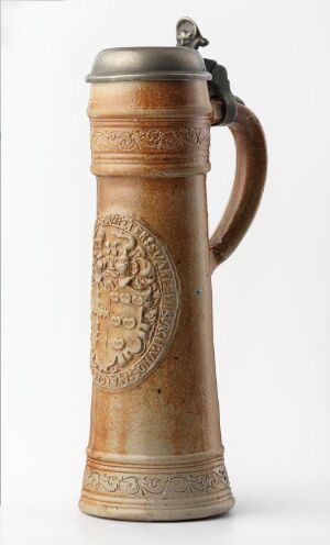  A traditional ceramic beer stein with a hinged pewter lid, featuring a central embossed emblem and decorative elements, in warm beige tones.