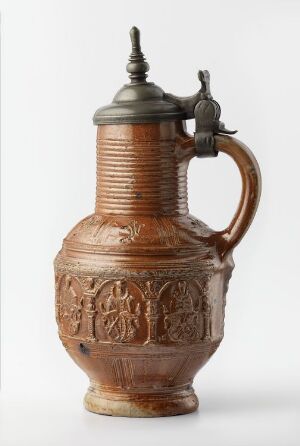  A traditional ceramic jug with a pewter lid, featuring a terra cotta color and decorated with relief patterns of stylized floral motifs and horizontal ribbing on the neck. The jug exhibits a robust handle and a slightly flaring base and is set against a plain light background.