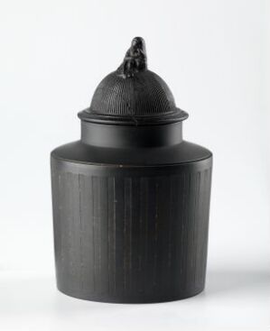  A dark grey, matte cylindrical container with a domed lid featuring a decorative knob, set against an off-white background.