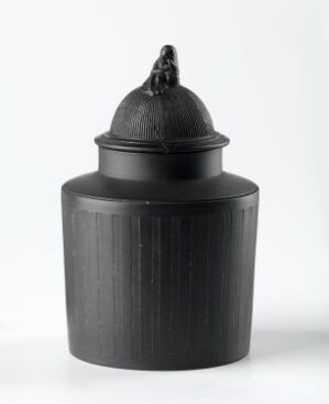  A dark gray cylindrical container with a dome-like lid and vertical ridges, featuring a knob on top, against a light gray background. The artist's name and title are unknown.