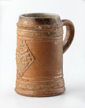  A cylindrical terracotta mug with a single handle, featuring a dark rim and a large incised triangular pattern filled with crisscrossing lines on the front, against an off-white background.